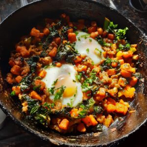 A cast iron skillet is shown. It is filled with a sweet potato and sausage hash, with two fried eggs sitting within the mixture.